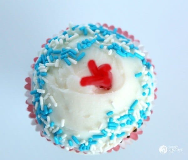 Looking down on one of the Buttermilk Firecracker Cupcakes, white frosting with blue and white sprinkles around and red licorice sprouting up from the middle like a firecracker. 