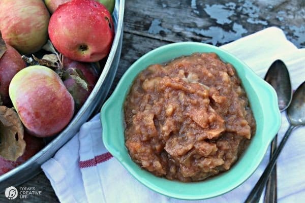 Vintage blue green bowl filled with chunky crockpot slow cooker applesauce. Large bucket filled with apples next to applesauce.