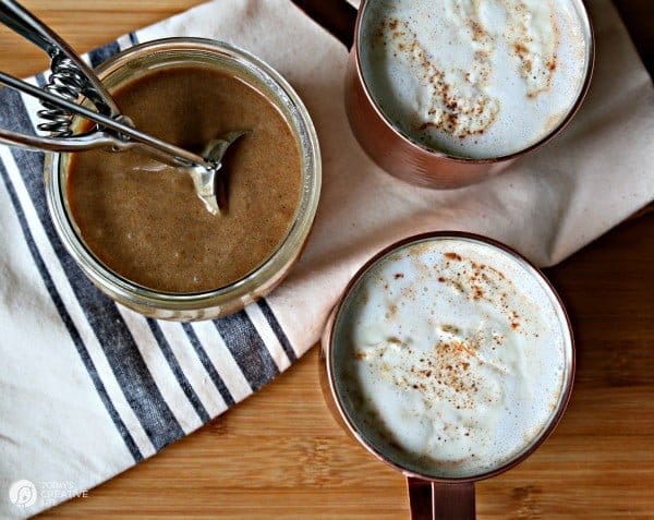 Jar filled with hot buttered rum mix with 2 mugs filled, nutmeg sprinkled on top.