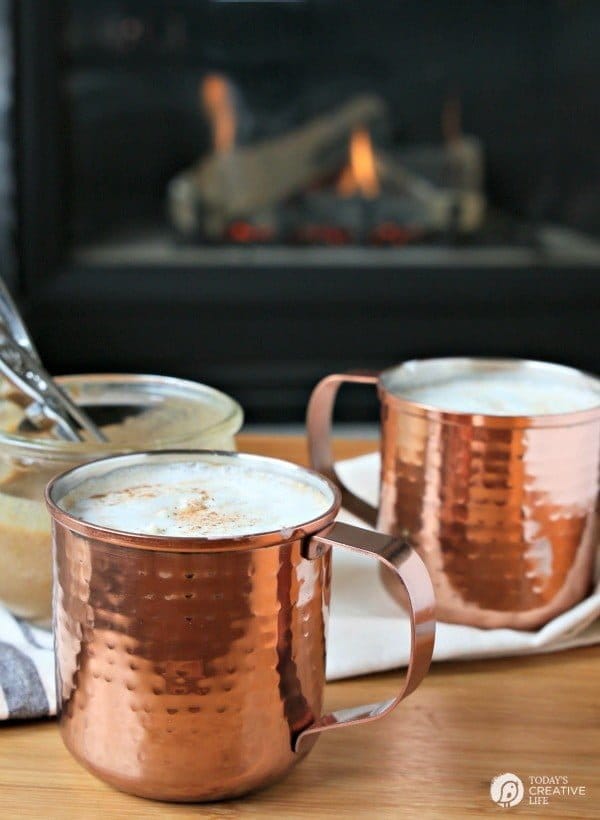 2 copper mugs filled with hot buttered rums next to a fire. 