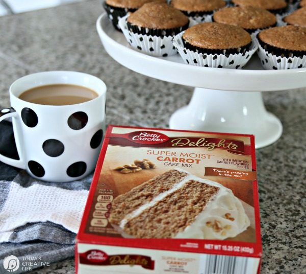 Muffins on a white cake plate, a cup of coffee in a black and white polka dot mug and a box of cake mix.