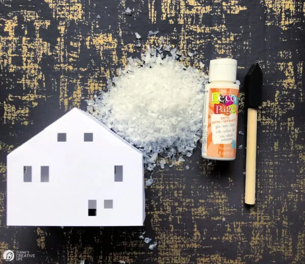 Paper house with a bottle of mod podge, a craft brush and faux snow.