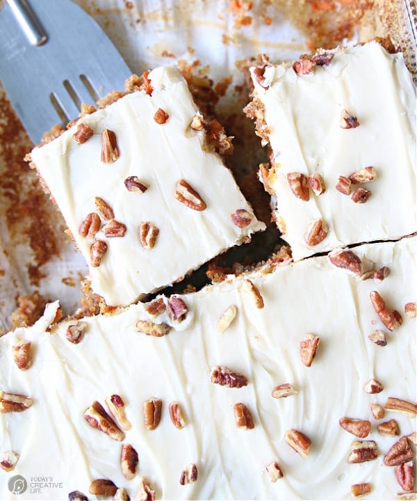 Carrot Cake Recipe with pecan sprinkled on cream cheese frosting in a cake pan being cut and served. 