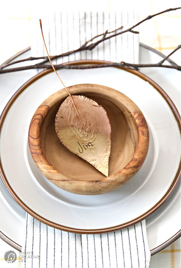 Gold painted leaf inside a small wooden bowl that's placed on white plates. Ideas for Thanksgiving Table Settings