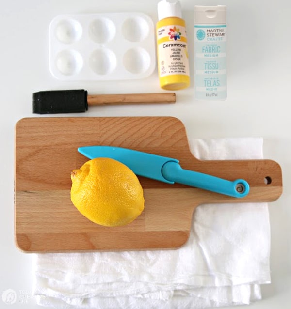 Supplies for making DIY Lemon Stamped Tea Towels: Lemon, knife, acrylic paint, fabric medium, craft brush on cutting board.