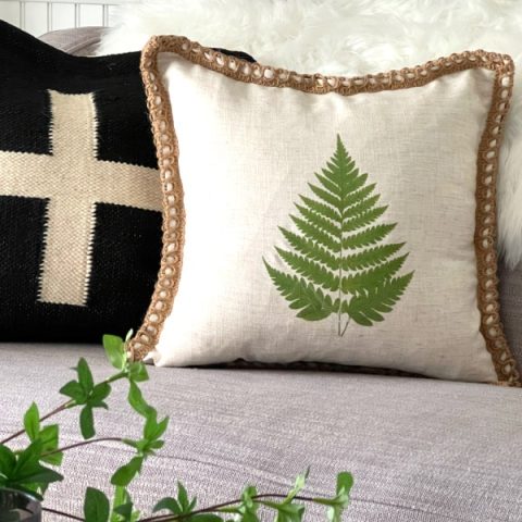 Two pillows on sofa. One pillow with a green fern. The other pillow is black with white cross.