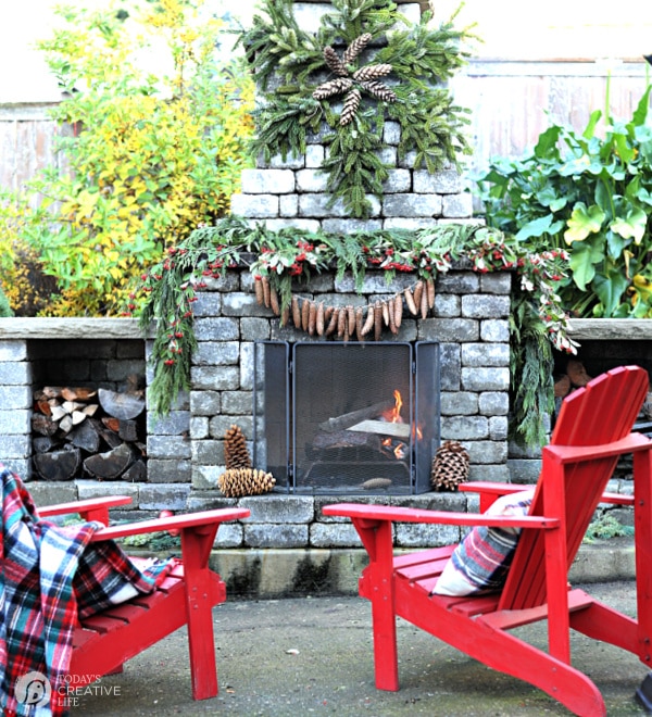 Outdoor holiday decorating ideas for Christmas. Red Adirondack chairs sitting next to an outdoor fireplace that's decorated for Christmas. 