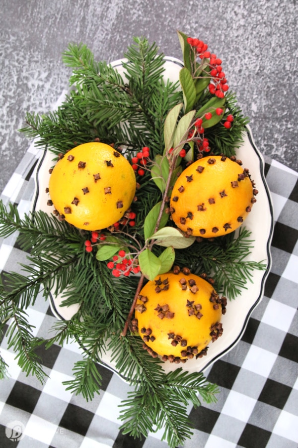 3 oranges with cloves on a plate decorated with Christmas greens. 